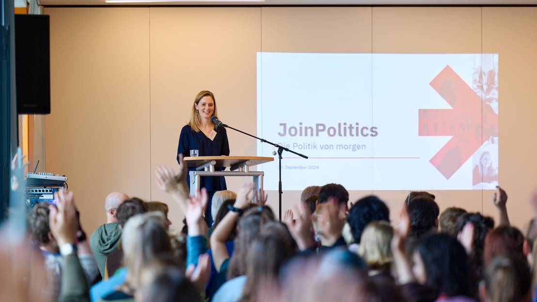 Caroline Weimann steht auf einer Bühne vor einer Präsentation mit dem Schriftzug "Join Politics" vor ihr heben einige im Publikum die Hand für Fragen