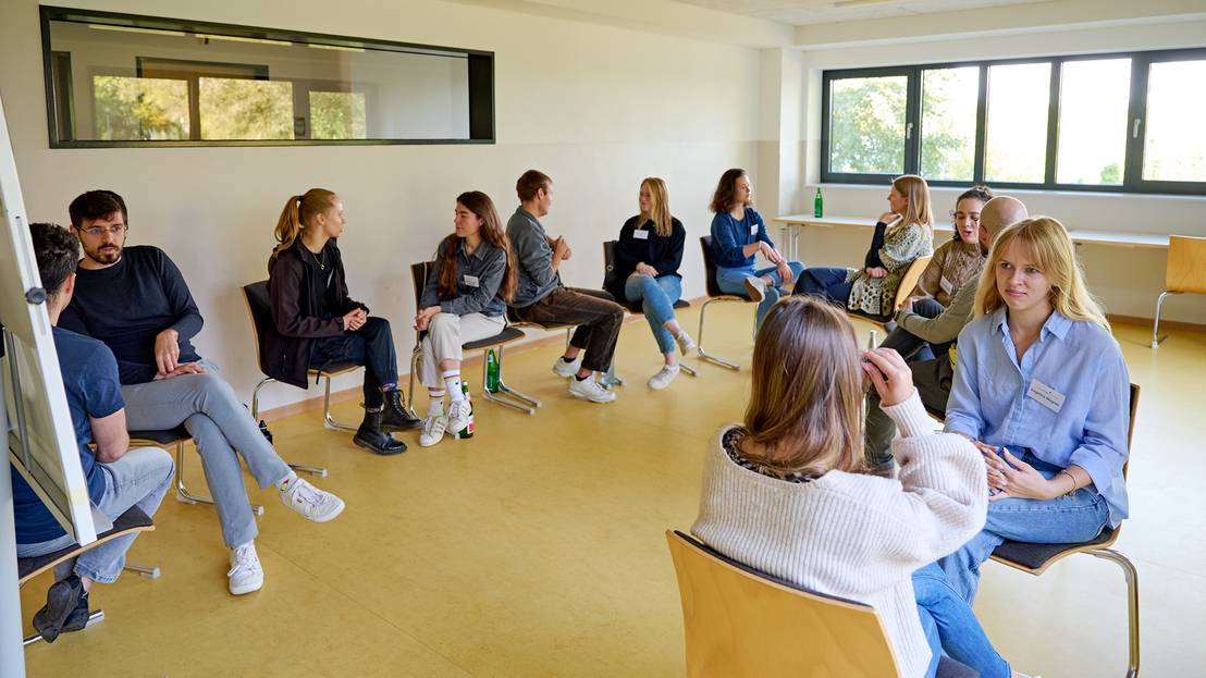 Workshopraum mit Stuhlkreis: Zwölf Teilnehmende sitzen in sechs Zweiergruppen einander zugewandt und diskutieren angeregt.