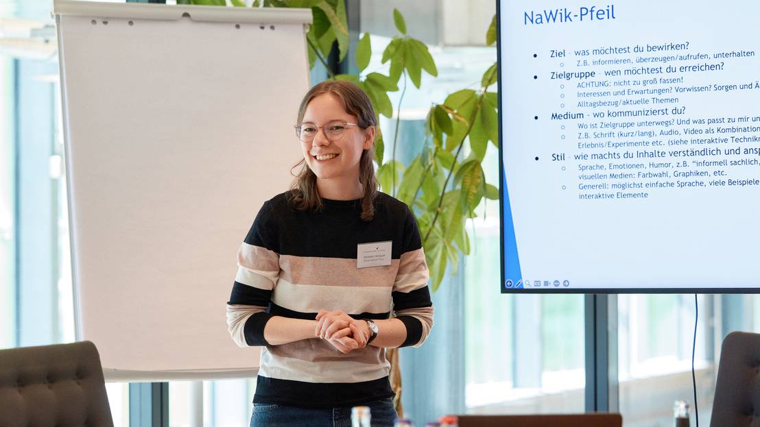 Stefanie Henauer hält entspannt lachend einen Vortrag umrahmt von Flipchart und Powerpoint Präsentation