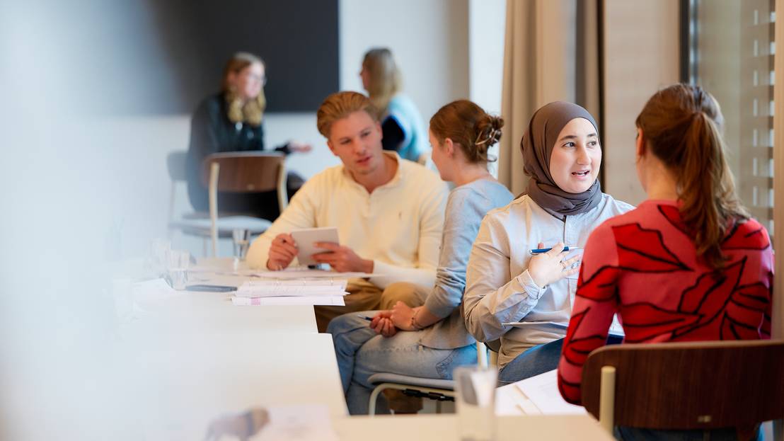 In einem Workshop-Raum sitzen drei Zweiergruppen an Tischen und sprechen miteinander. Die Teilnehmenden sind in angeregte Diskussionen vertieft.